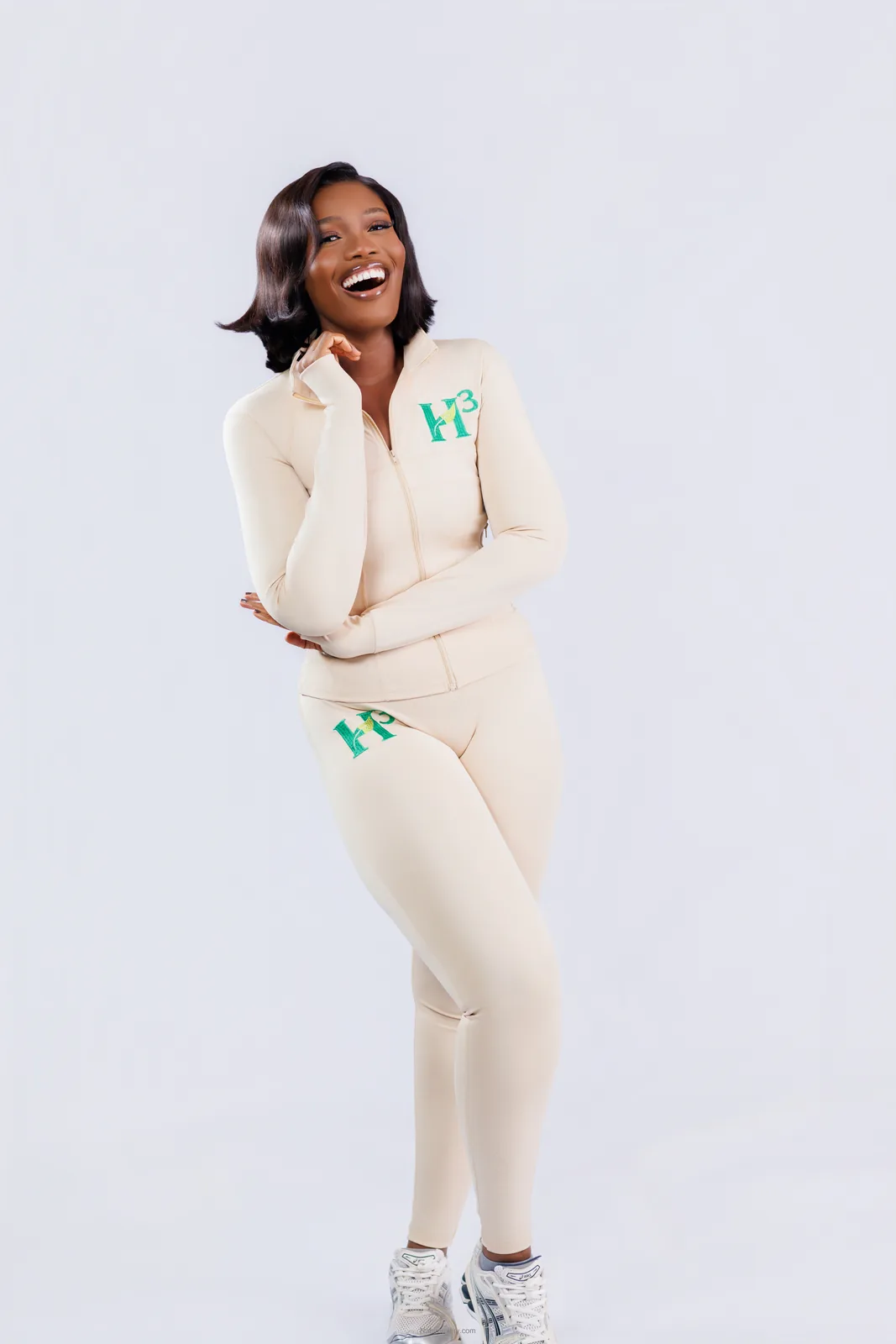 A woman standing and laughing in a cream H³ tracksuit, hand raised to her chin, against a pale studio backdrop.